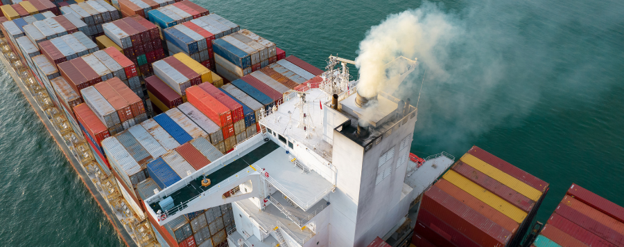 image of a large ship filled with metal shipping containers. Smoke is coming out of the smoke stack on the ship.